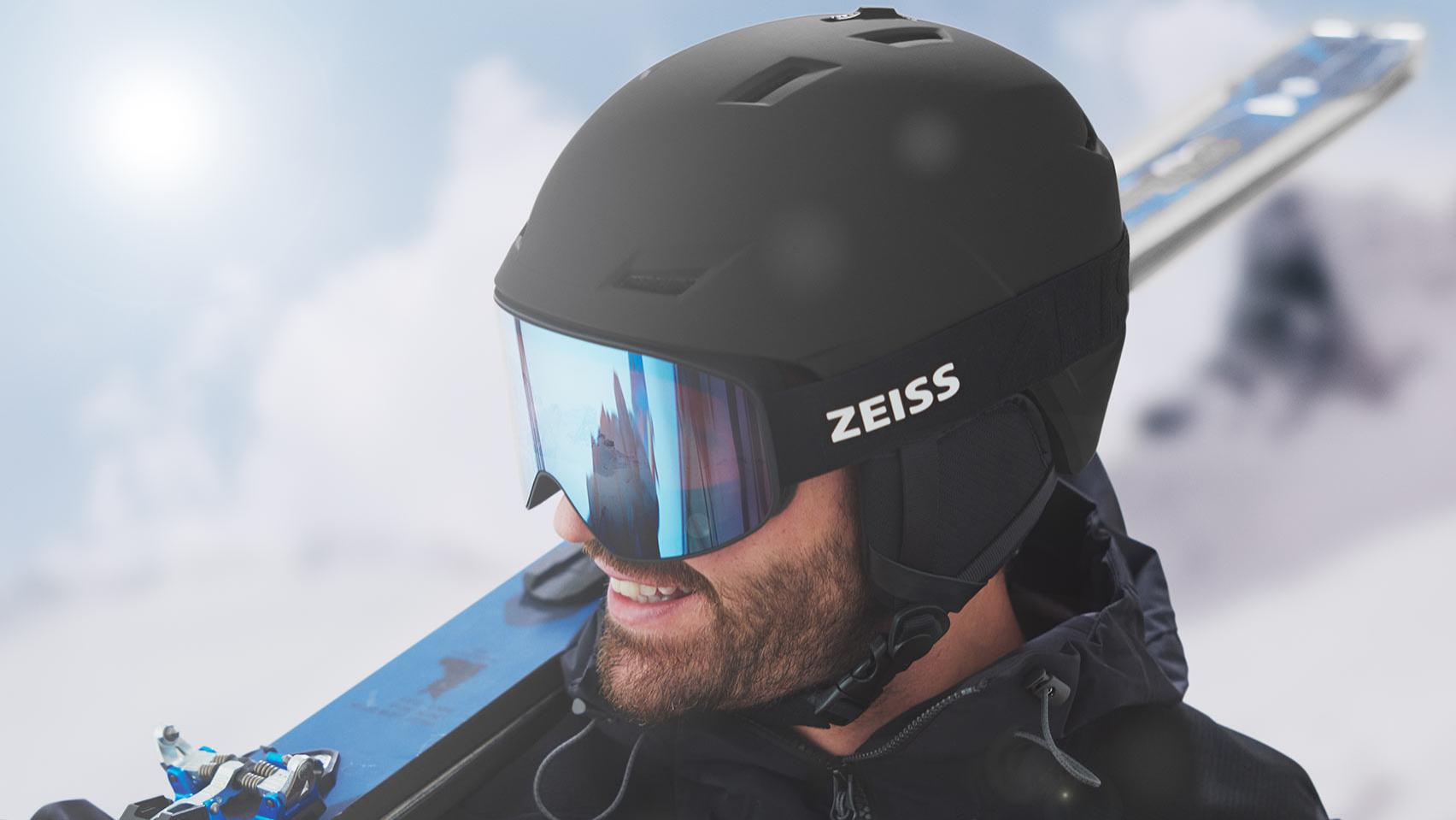 Un homme porte un casque noir et des lunettes de ski ZEISS à miroir bleu, avec des skis sur l'épaule droite.