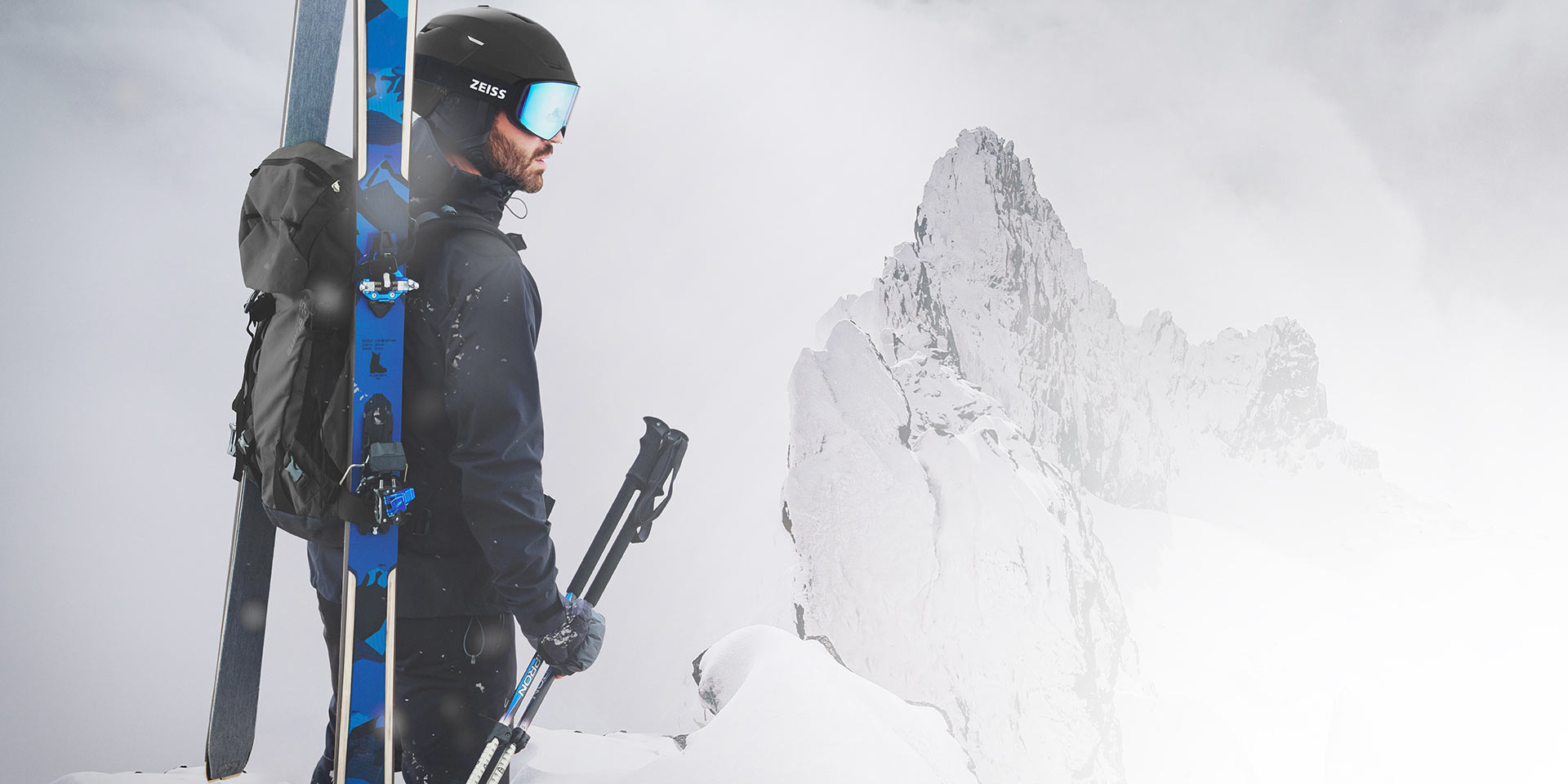 Un homme porte un casque noir avec des lunettes de ski ZEISS à miroir bleu, des skis et un sac à dos sur le dos et tient les bâtons de ski dans sa main droite. Il se tient au sommet d'une montagne.