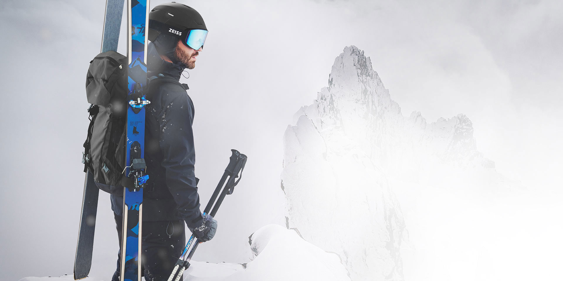 Un homme porte un casque noir avec des lunettes de ski ZEISS à miroir bleu, des skis et un sac à dos sur le dos et tient les bâtons de ski dans sa main droite. Il se tient au sommet d'une montagne.