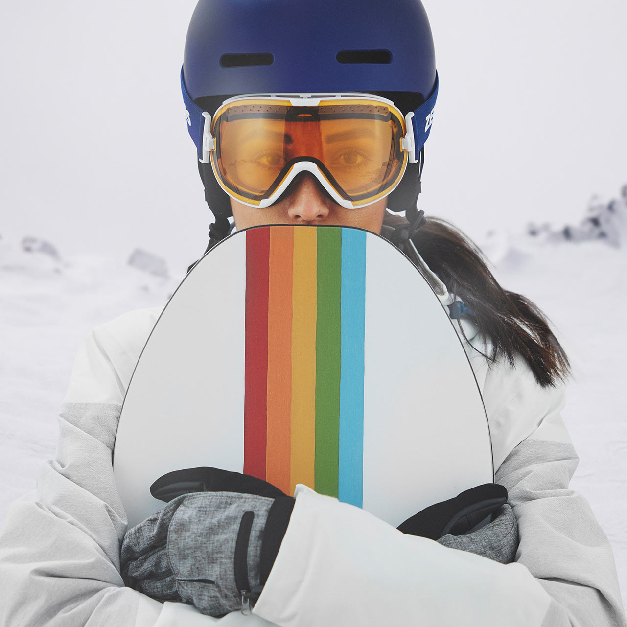 Une femme porte un casque bleu et des lunettes de ski ZEISS à miroir bleu, tenant un snowboard coloré dans sa main droite. Montagnes enneigées en arrière-plan.