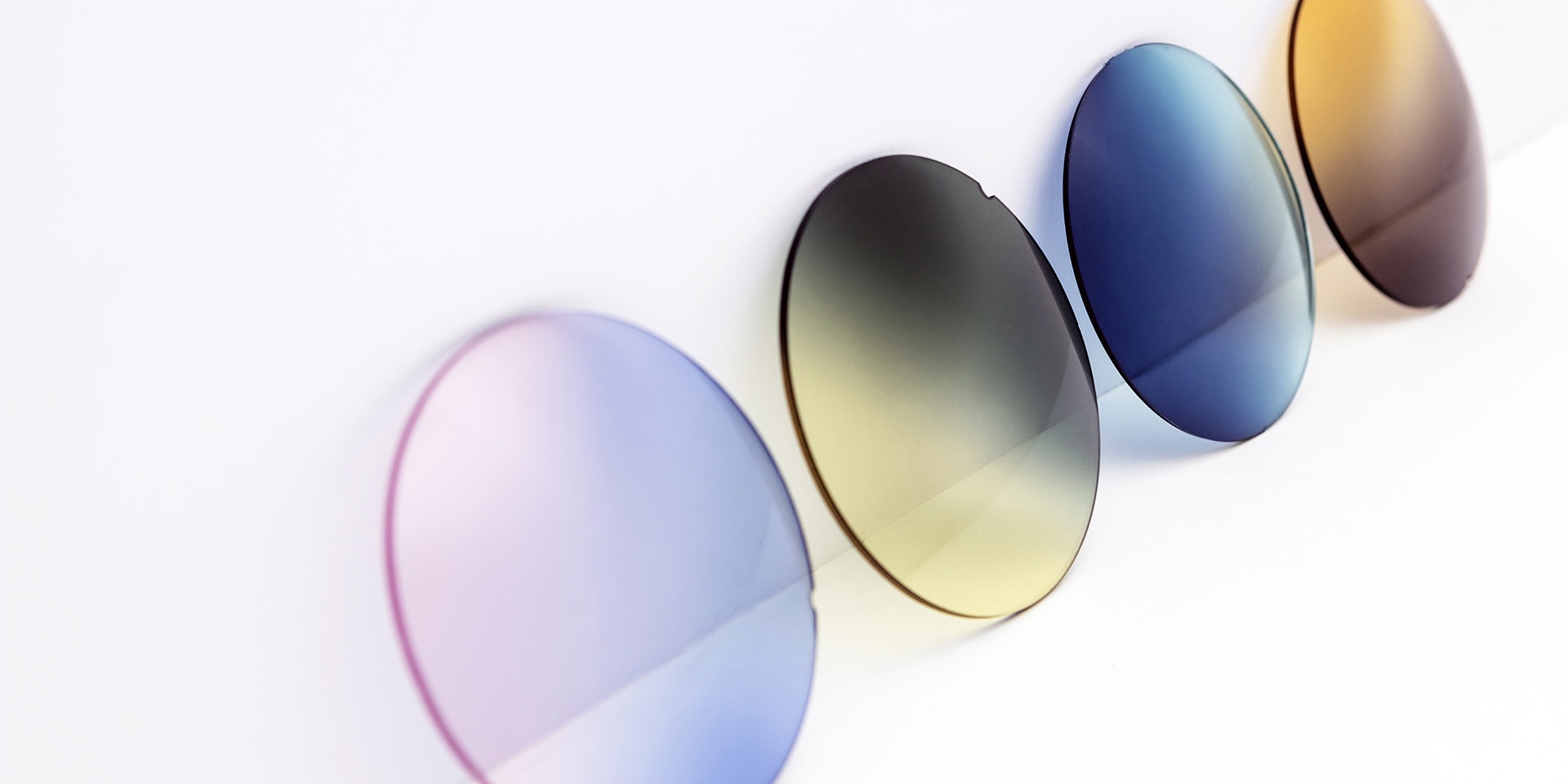 Des verres de lunettes de soleil de différentes couleurs appuyés contre une surface blanche : des dégradés rose-violet, jaune-gris, bleu et marron.