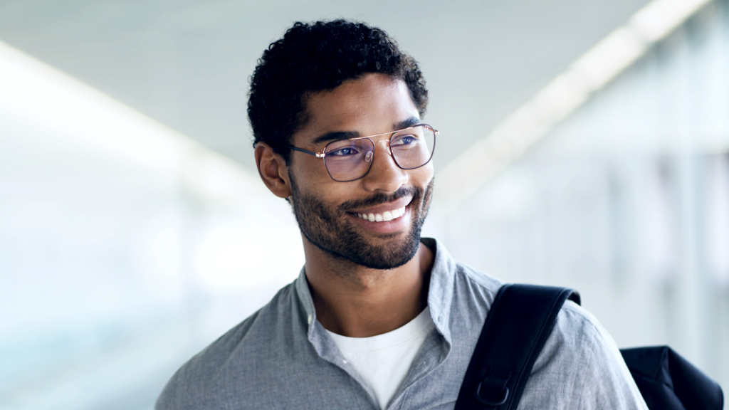 Un jeune homme sourit avec un sac à dos sur l’épaule, il porte des verres ZEISS Digital SmartLife et regarde sur le côté.